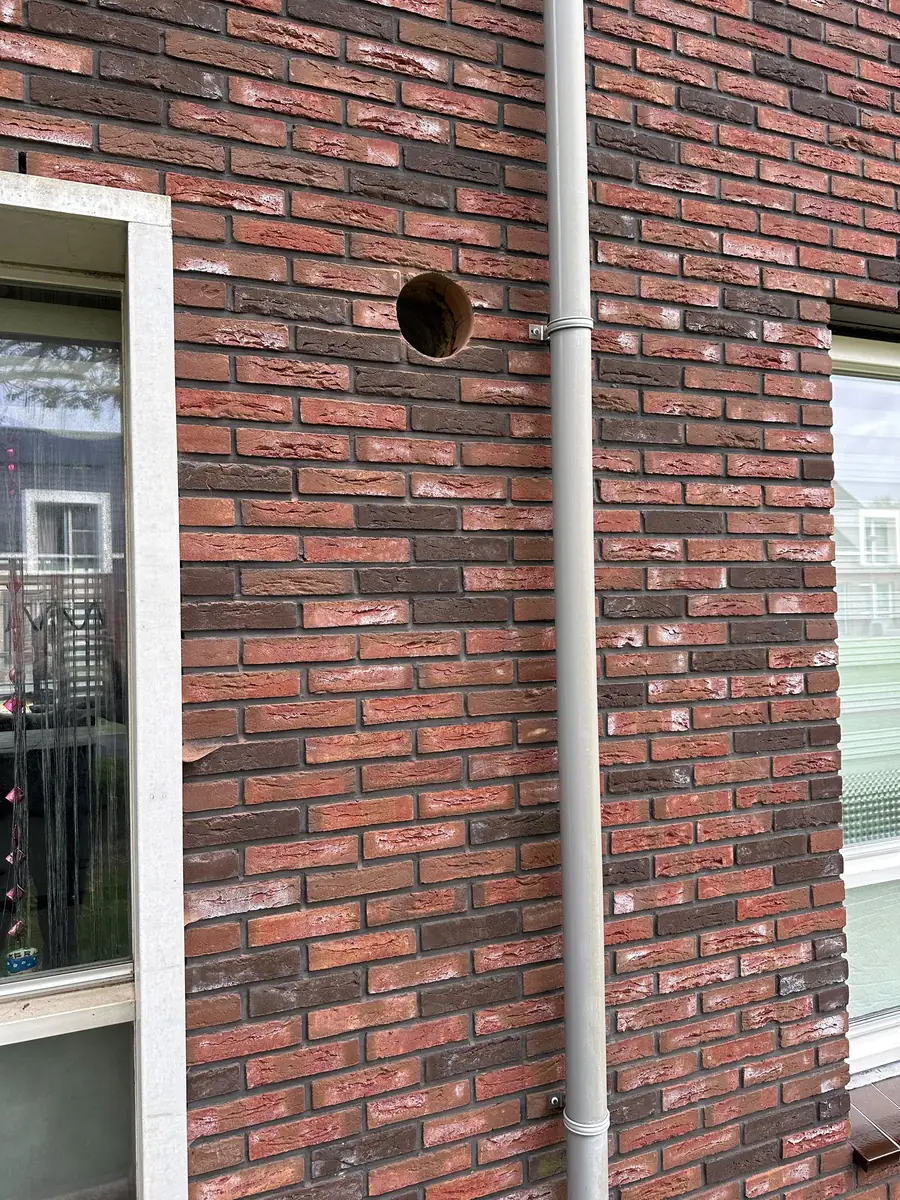 Afzuigkap gat boren Venray &mdash; Afzuigkapgat in een roodbonte gevel tussen twee kozijnen, naast een ronde regenpijp
