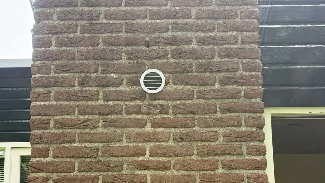 Rond wit gevelrooster in een bakstenen buitenmuur, geplaatst in een kernboring voor de ventilatie van de ruimte erachter