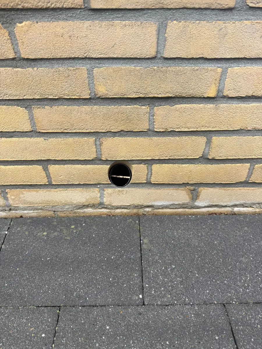 Kruipruimte ventilatie 's-Hertogenbosch &mdash; Rond geboord ventilatiegat met koker in een gele bakstenen gevel, vlak boven de tegels