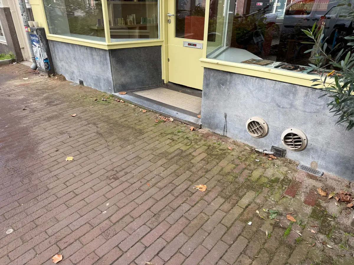 Kruipruimte ventilatie Roosendaal &mdash; Twee ronde ventilatieroosters in de grijze plint onder een etalage, boven de klinkers