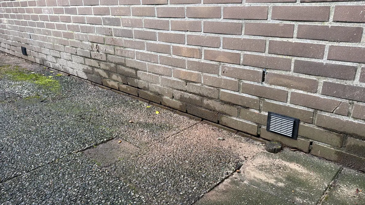 Kruipruimte ventilatie Roosendaal &mdash; Zwart ventilatierooster laag in een bruine bakstenen gevel, boven grijze betontegels