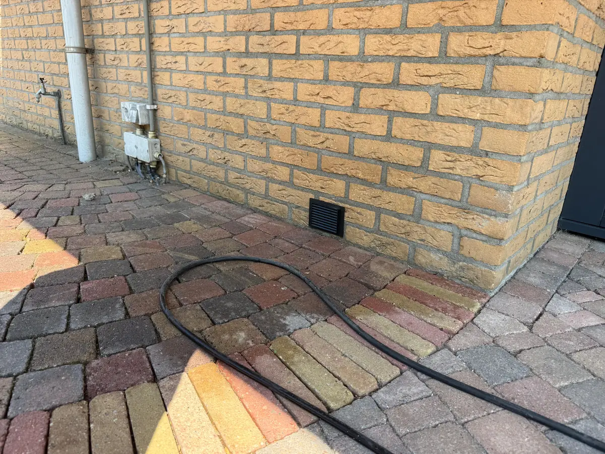 Kruipruimte ventilatie 's-Hertogenbosch &mdash; Ventilatierooster laag in een gele bakstenen gevel, met een tuinslang op de klinkers