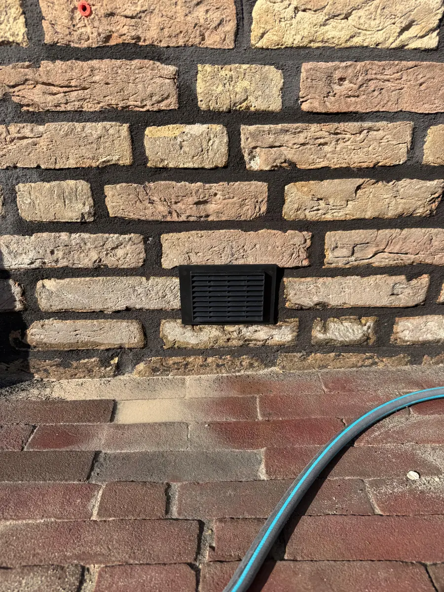 Kruipruimte ventilatie Roermond &mdash; Zwart ventilatierooster in een gele gevel met donkere voegen, met de waterslang ernaast