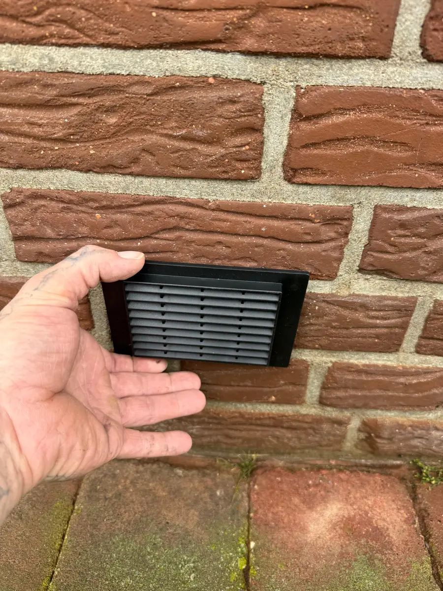 Een zwart ventilatierooster wordt in een bakstenen gevel geplaatst voor de kruipruimte