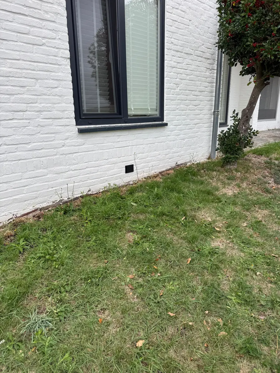 Kruipruimte ventilatie Oosterhout &mdash; Zwart ventilatierooster in een witgeschilderde bakstenen gevel, net boven het gras