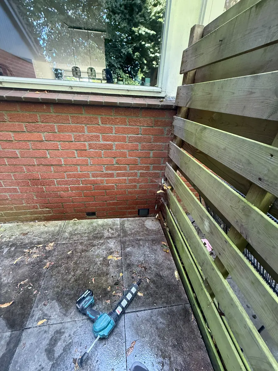 Kruipruimte ventilatie Kerkrade &mdash; Twee ventilatieroosters onderaan een rode bakstenen gevel, in de hoek bij een houten schutting