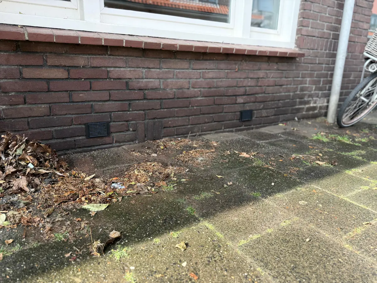 Kruipruimte ventilatie Heerlen &mdash; Ventilatieroosters onderaan een donkere bakstenen gevel, boven tegels met blad