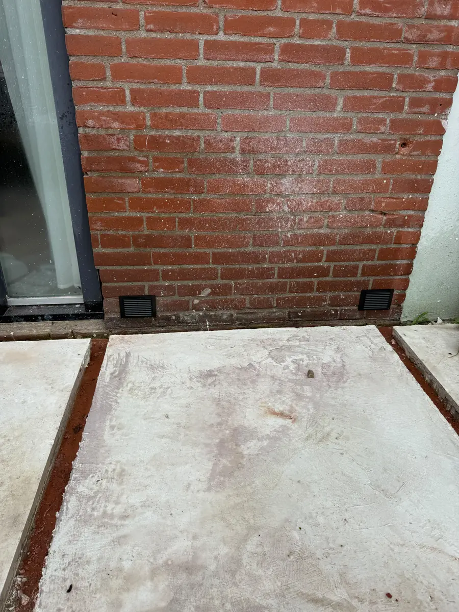 Kruipruimte ventilatie Helmond &mdash; Twee ventilatieroosters van de kruipruimte in een rode gevel, net boven betonnen terrasplaten