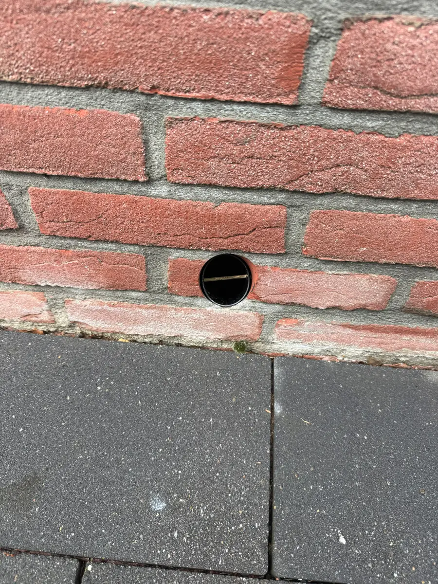 Kruipruimte ventilatie Helmond &mdash; Rond ventilatiegat met koker in een rode gevel, vlak boven grijze tegels
