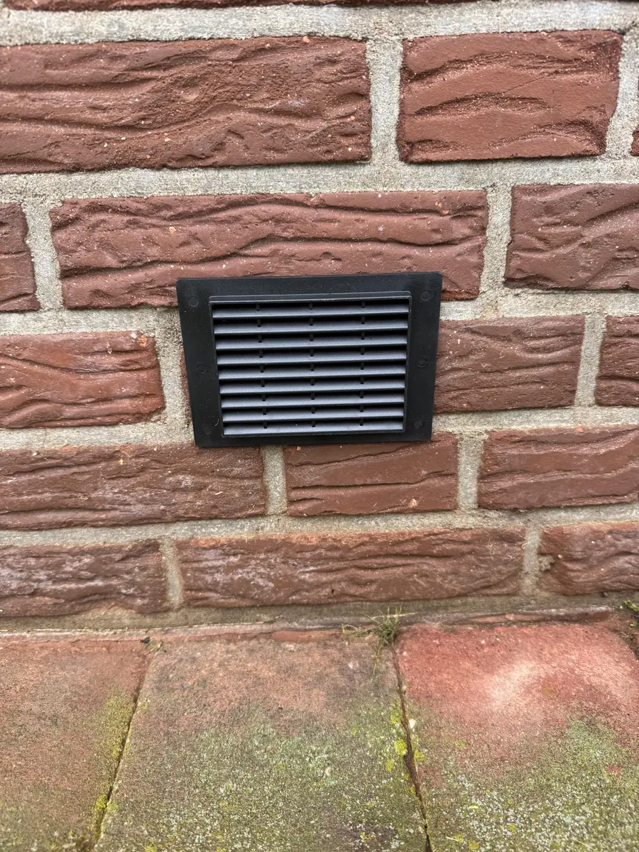 Kruipruimte ventilatie Tilburg &mdash; Zwart ventilatierooster van de kruipruimte in een rode bakstenen gevel, net boven de tegels