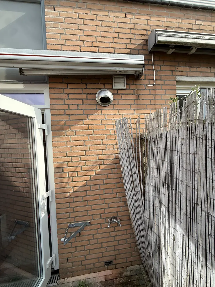 Afzuigkap gat boren Veldhoven &mdash; Rvs-buitenkap van een afzuigkap in een gele gevel aan de tuinzijde, naast een ventilatierooster