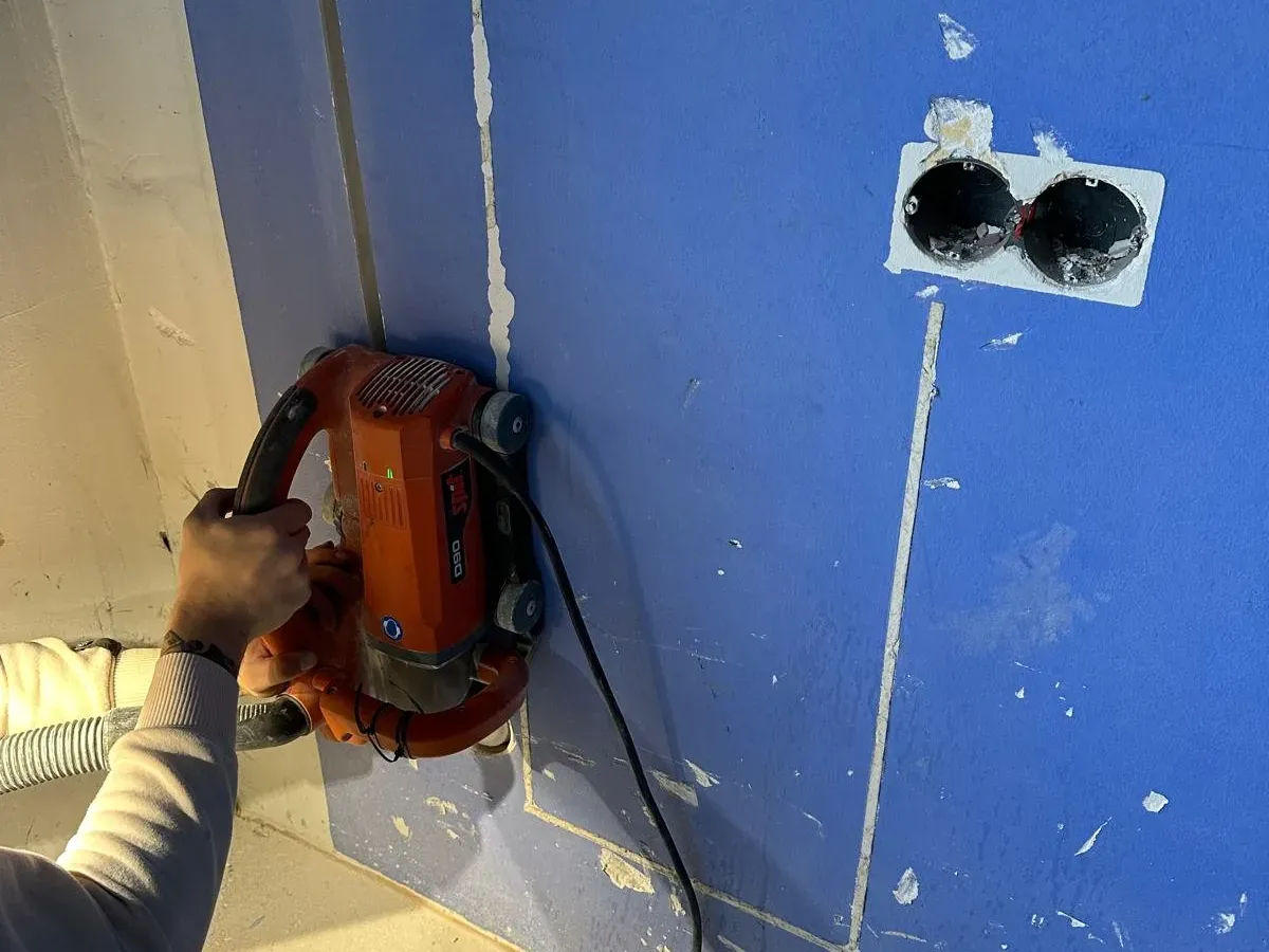 Sleuvenfrees in actie op een blauwe wand, met de stofzuigerslang aan de machine en een dubbel stopcontact